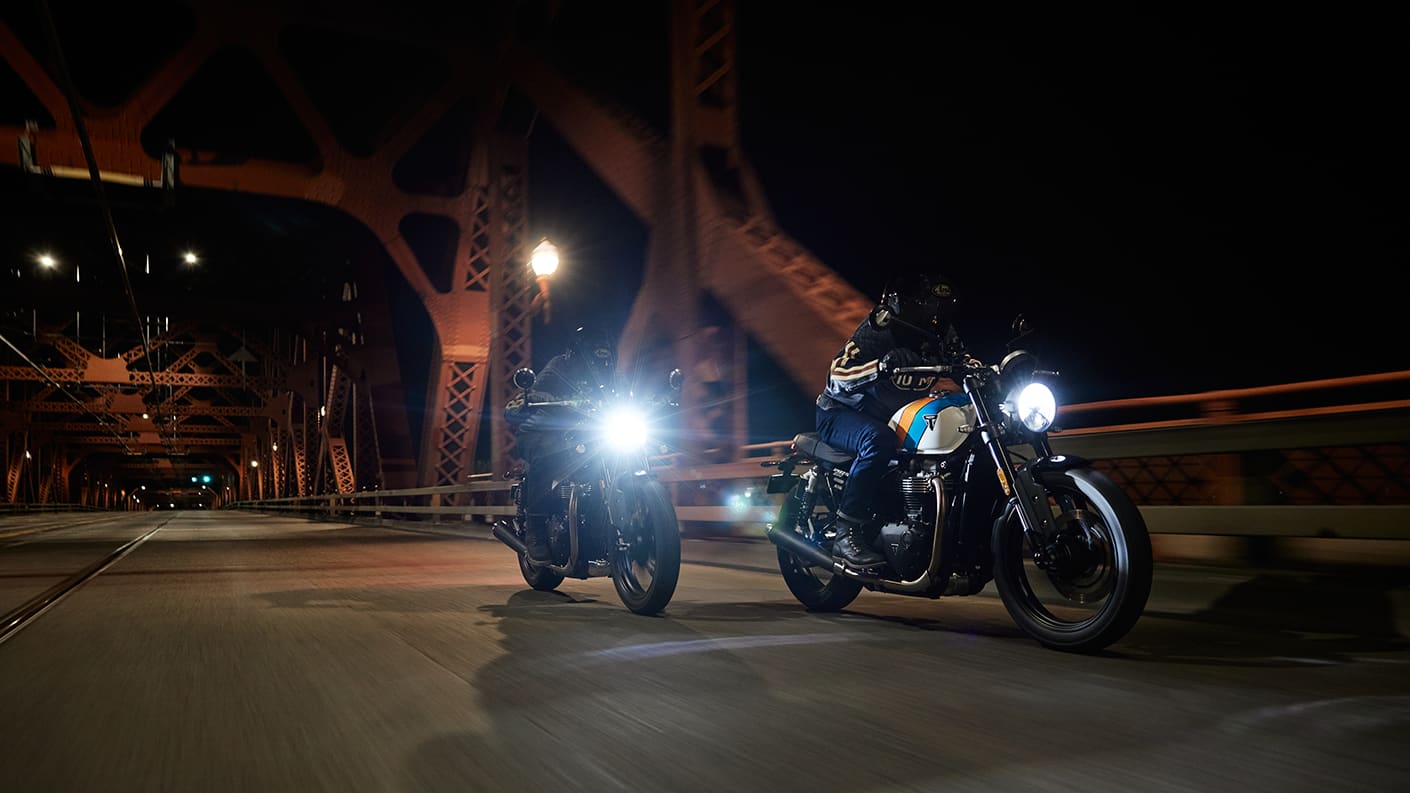 Two Triumph Speed Twin 900s riding towards the camera on urban bridge at night